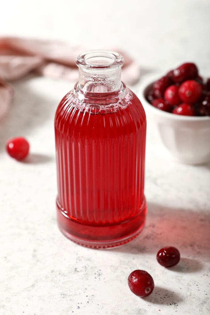 A ribbed glass jar of cranberry simple syrup with a bowl of cranberries and a pink linen on marble