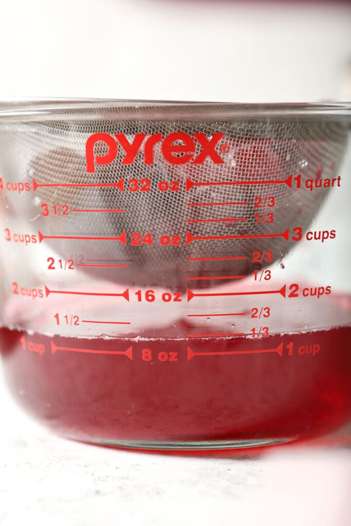 A strainer sits above a glass measuring cup straining out cranberries from a red liquid