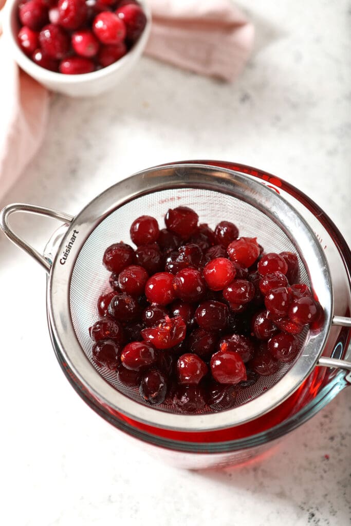 Cooked cranberries in a fine mesh sieve above a liquid measuring cup on marble