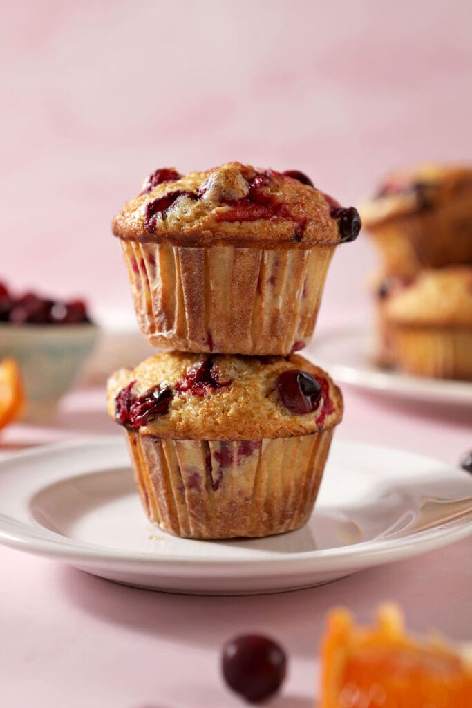 A stack of two Cranberry Muffins on a white plate, surrounded by cranberries and orange slices