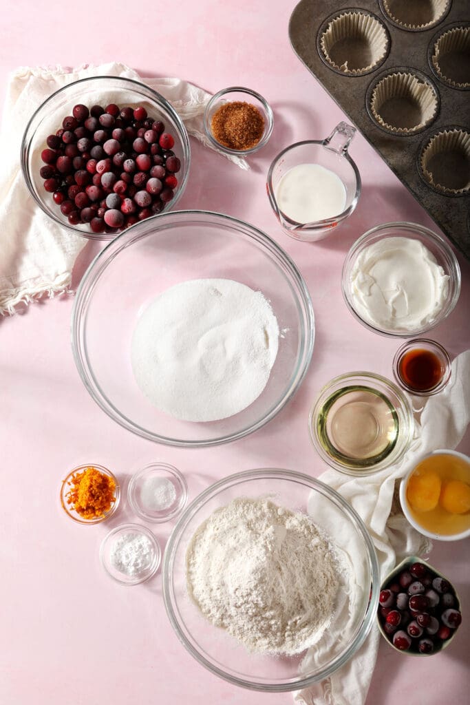 Ingredients to make muffins with cranberries in bowls on a pink countertop