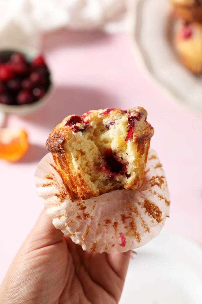 A hand holds a bitten-into Fresh Cranberry Muffin above a pink surface with a bowl of cranberries and orange slices