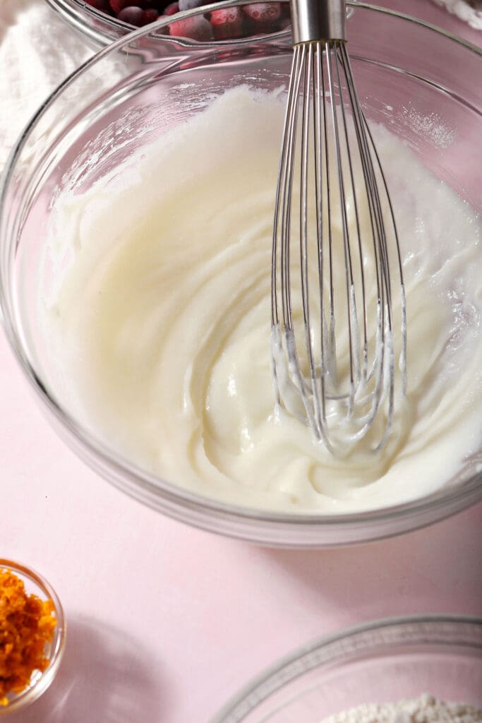 A whisk sits in a bowl with a sour cream-sugar mixture on a pink background