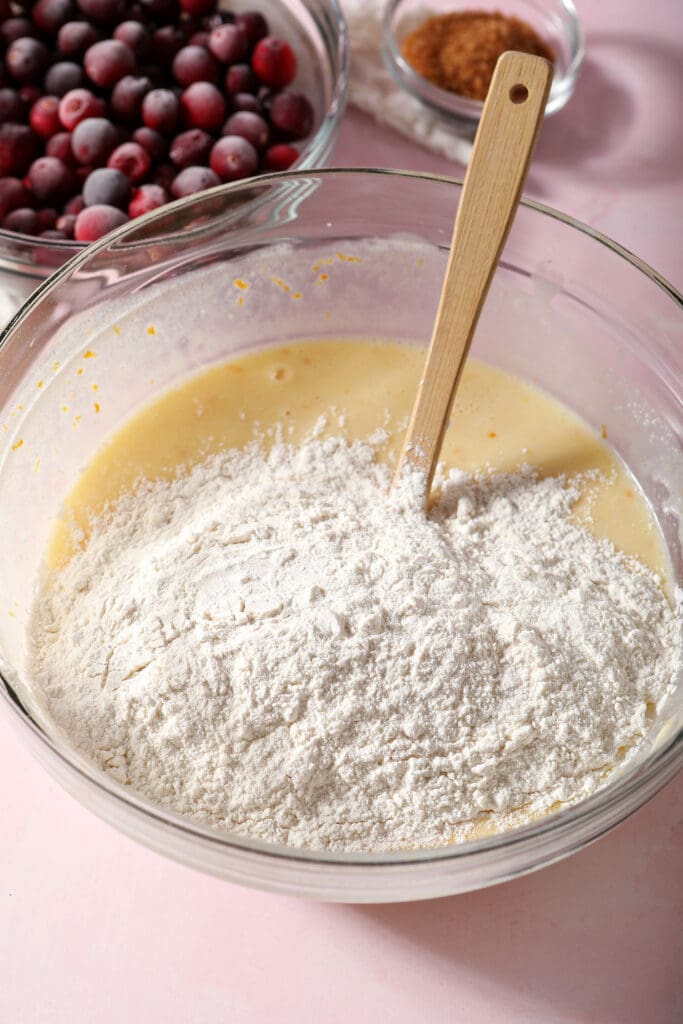 A flour mixture sits on top of wet ingredients in a bowl on a pink surface