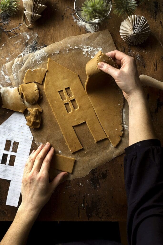 a set of hands cuts out a side of a gingerbread house from cookie dough on a wooden table