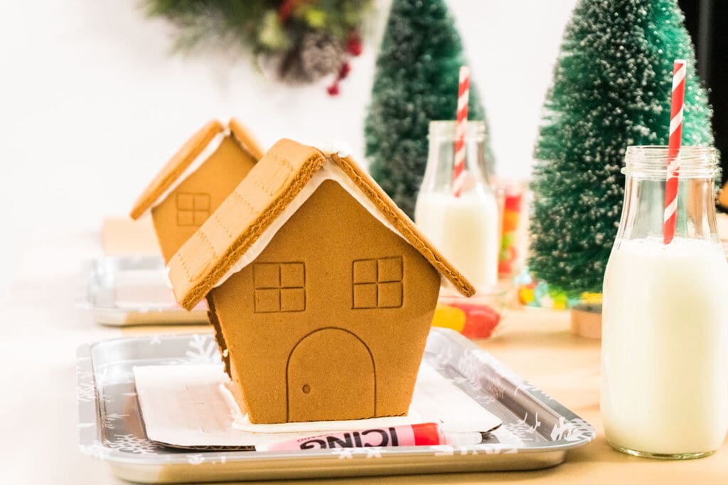 premade gingerbread houses on small sheet pans with icing sugar, glasses of milk, candies and more before kids decorate