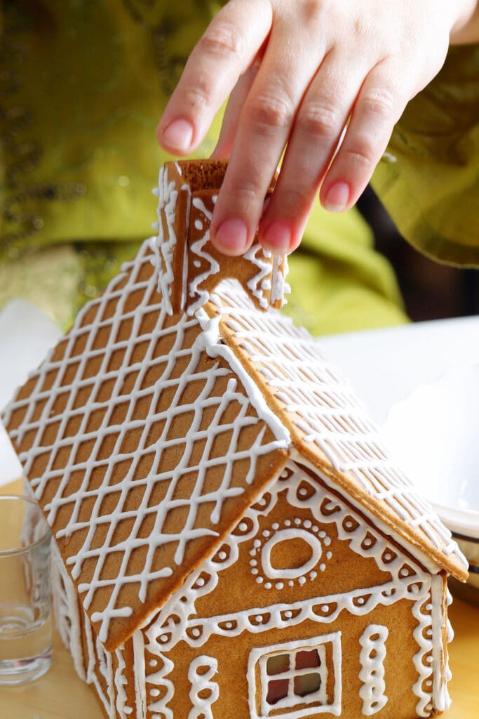 a hand presses a gingerbread chimney on top of a decorated gingerbread house with white icing as the decoration