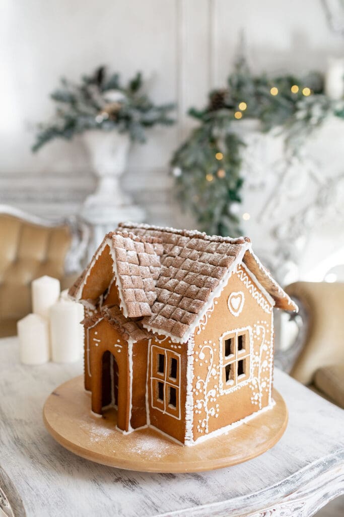 a decorated gingerbread house sits on a table in front of a tree and lit garland in a bright room