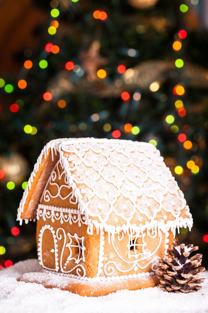 A frosted gingerbread house with white icing in front of a brightly lit christmas tree