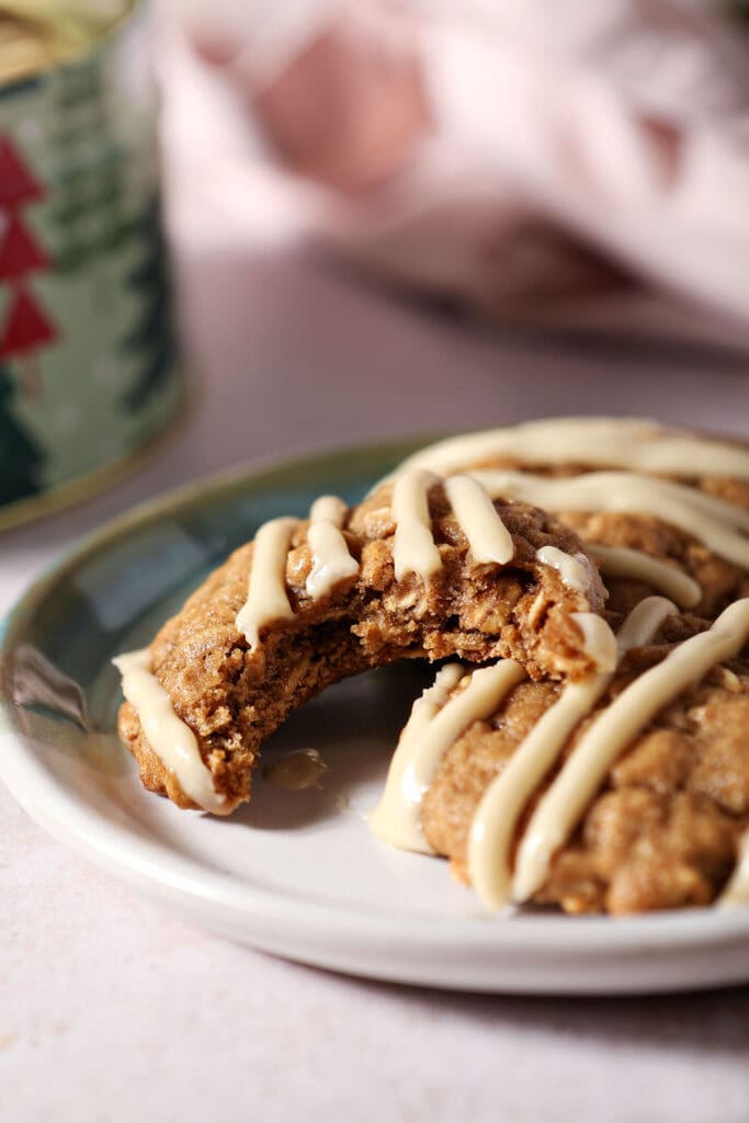 A bitten into Cinnamon Oatmeal Cookie on a plate with two others next to a Christmas tree cookie tin and a pink linen