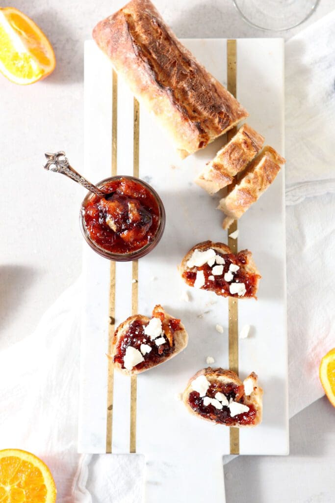 Overhead of three slices of crostini topped with cranberry compote next to a jar holding more compote and a half-sliced baguette on a marble cutting board