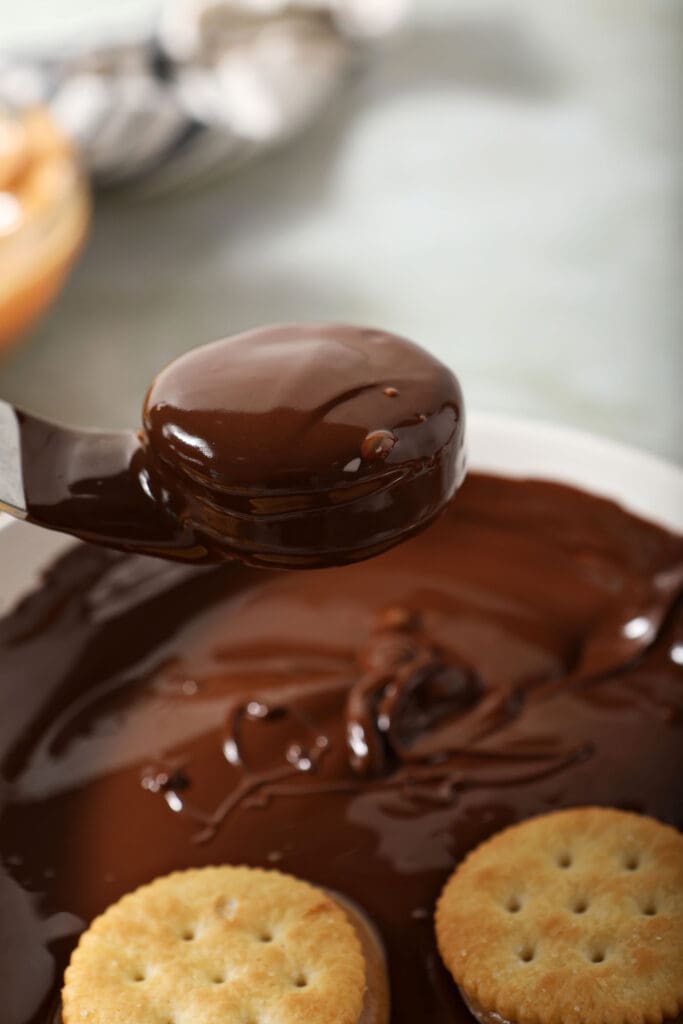 A chocolate covered ritz cracker cookie is lifted out of a bowl of melted chocolate with a fork