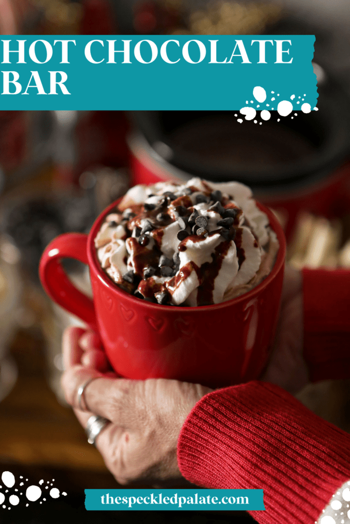 A woman holds a red mug of cocoa between her two hands above a hot chocolate bar with the text hot chocolate bar