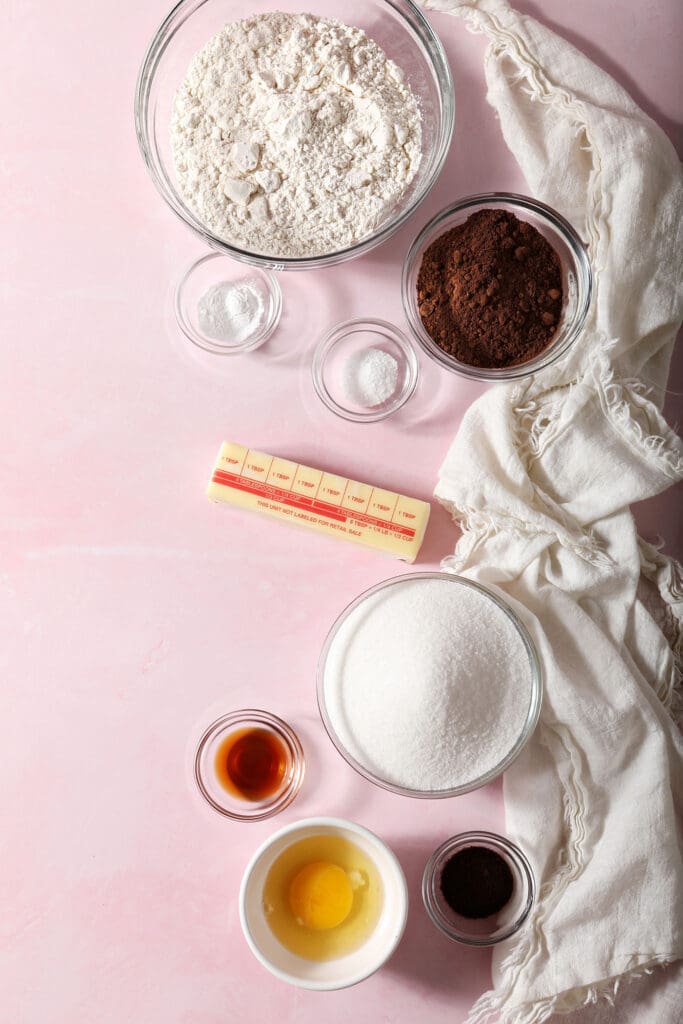 Ingredients to make chocolate cut out cookies in bowls on a pink surface with white linens