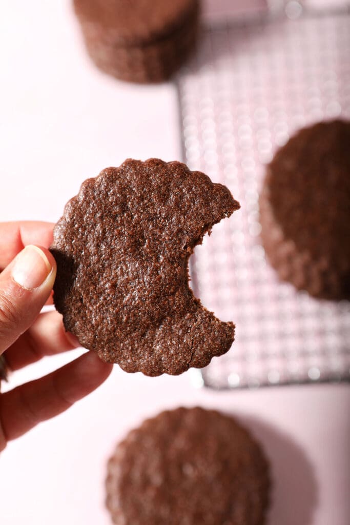 A hand holds a bitten-into chocolate sugar cookie above a pink surface holding more cookies