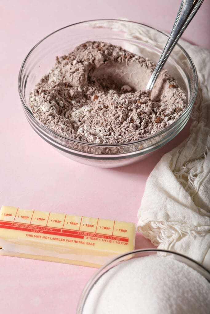 A mixing bowl with dry ingredients on a pink surface next to a stick of butter