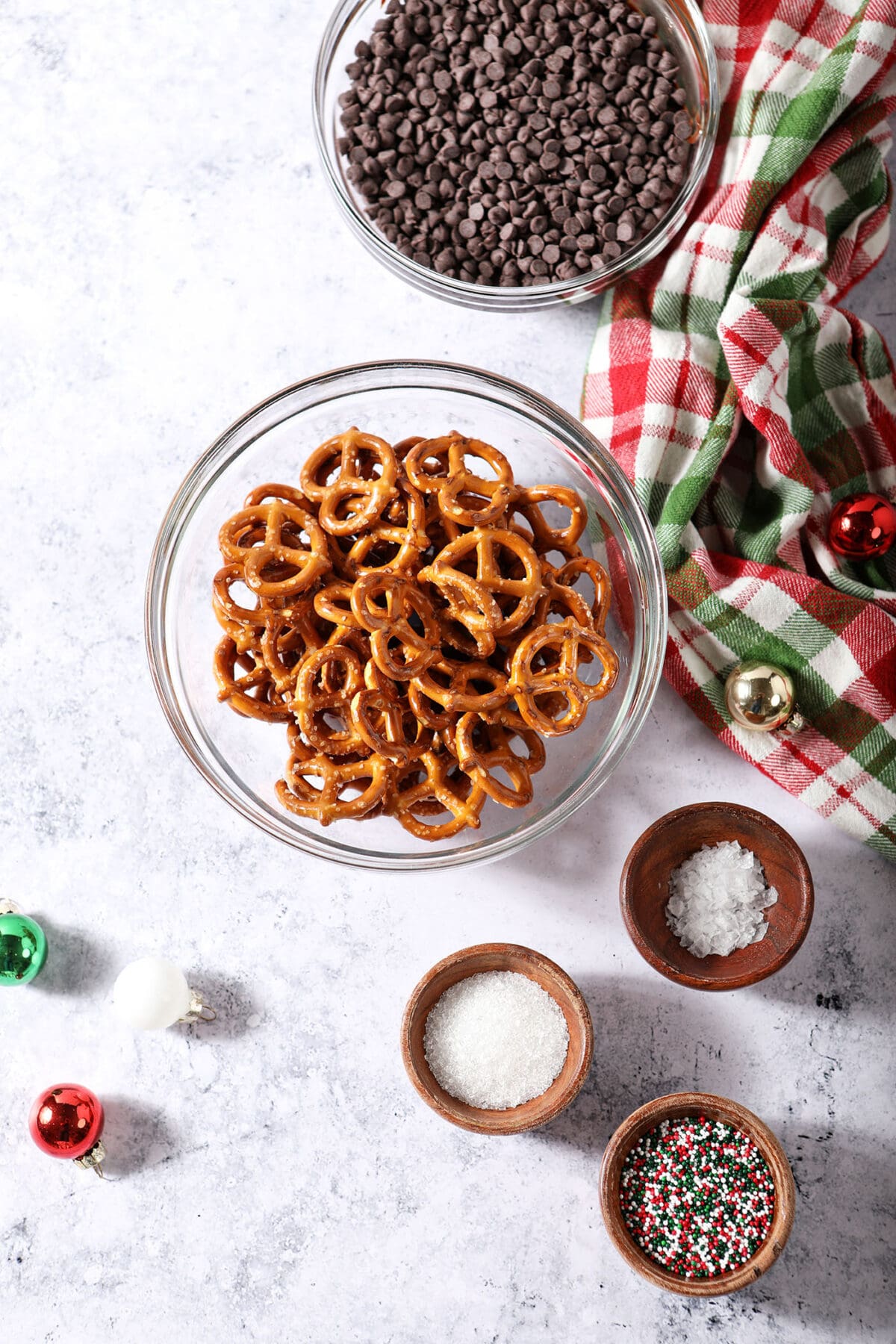 Ingredients to make chocolate dipped pretzels in bowls on a stone surface surrounded by ornaments and a red and green plaid linen