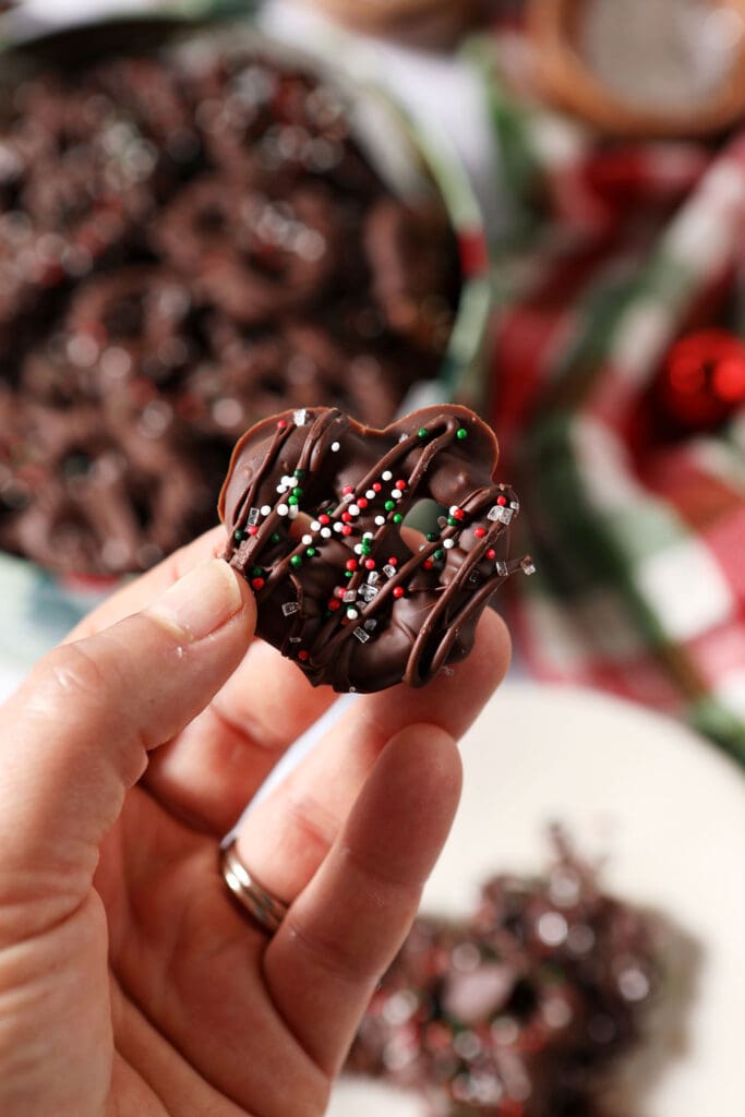 A hand holds a Christmas Chocolate Covered Pretzel above a tin and a plate of more pretzels