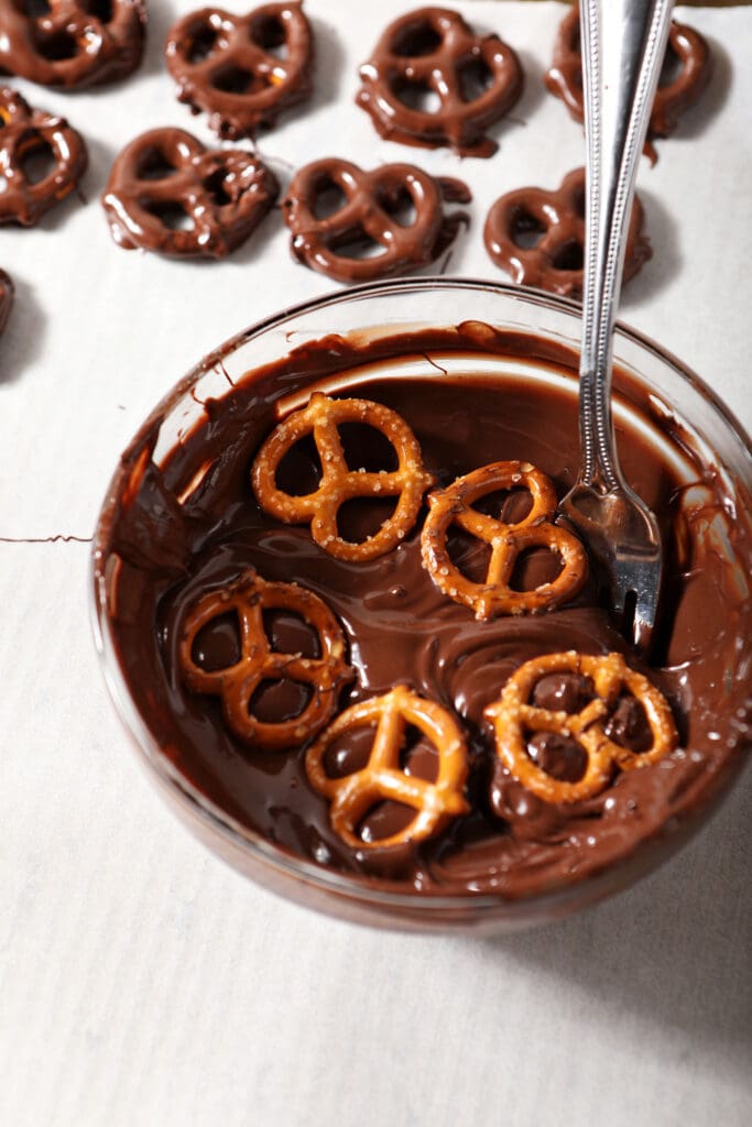 Five mini pretzels in a bowl of chocolate with a fork on a parchment-lined pan with chocolate dipped pretzels