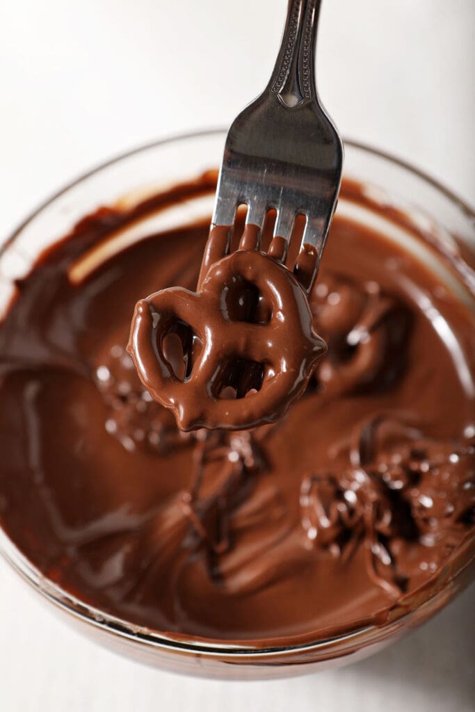 A fork lifts a chocolate covered pretzel out of a bowl of melted chocolate