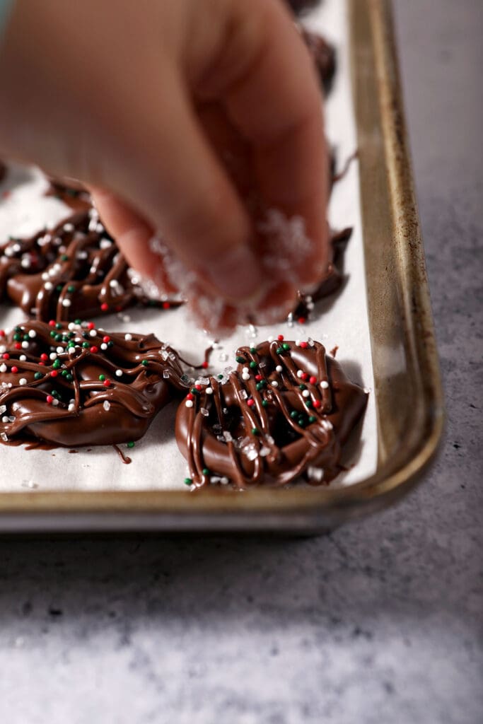 A child sprinkles sprinkles on top of chocolate dipped pretzels on a rimmed sheet pan