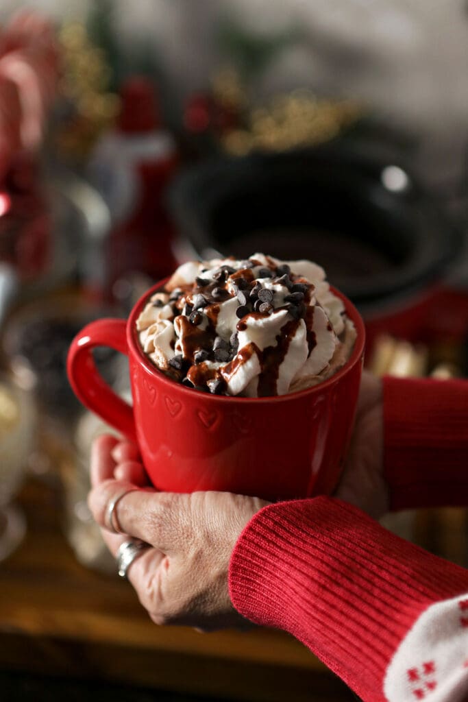 A woman holds a red mug of cocoa topped with whipped cream and chocolate sauce