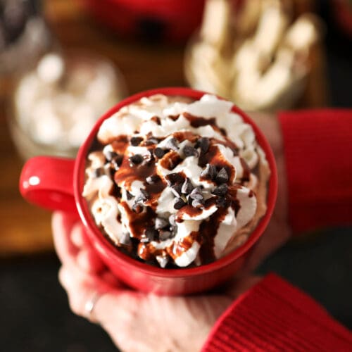 A woman holds a red mug of cocoa between her two hands above a hot chocolate bar