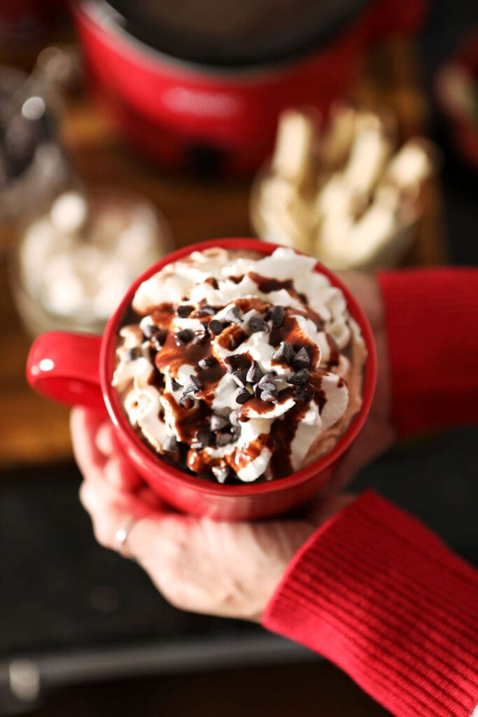 A woman holds a red mug of cocoa between her two hands above a hot chocolate bar