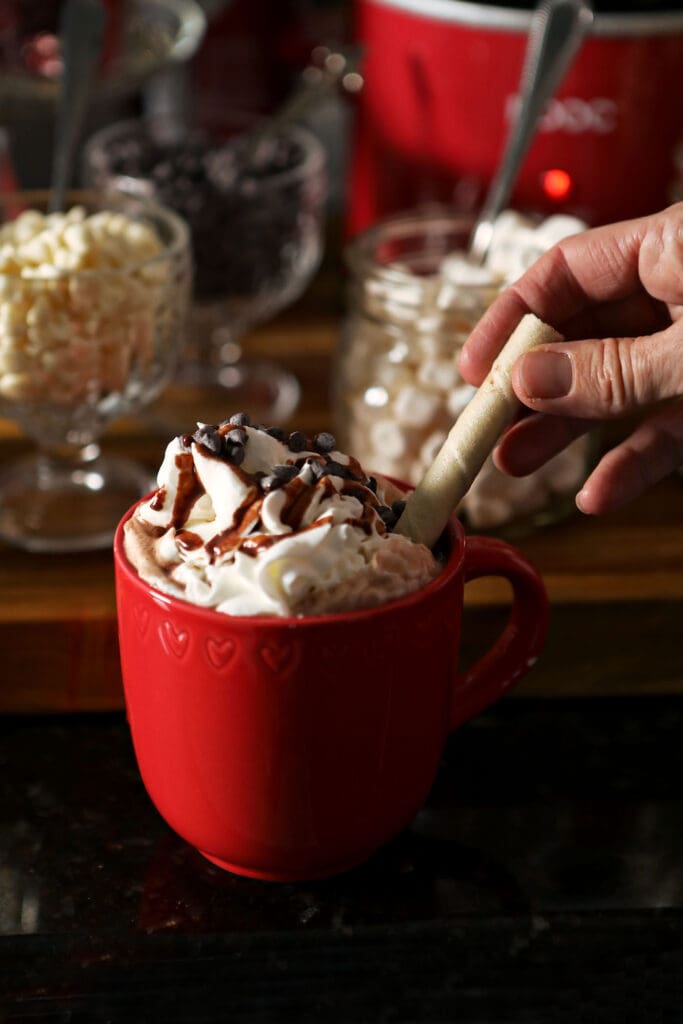 A hand sticks a rolled cookie into a whipped cream topped mug of cocoa