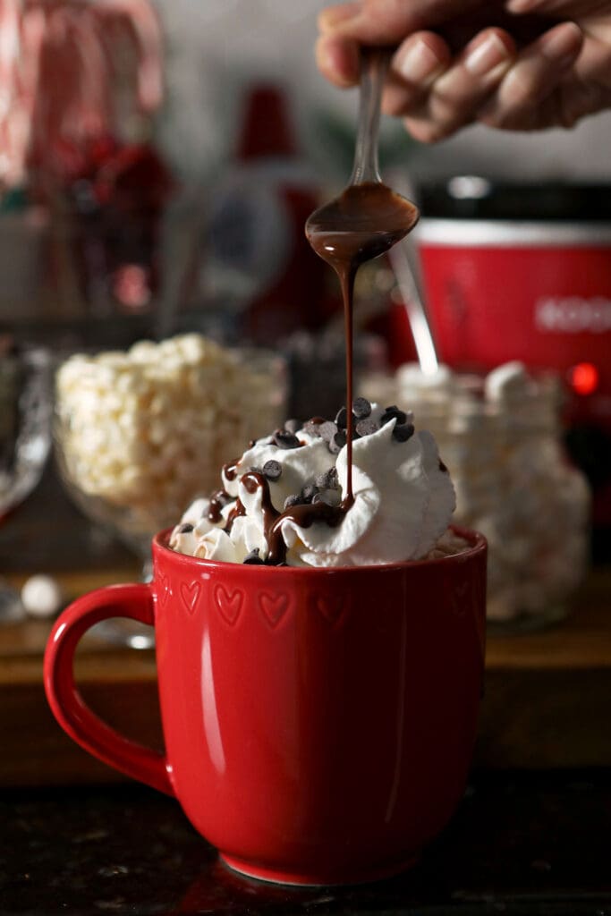 Chocolate syrup pours on top of a red mug of cocoa topped with whipped cream