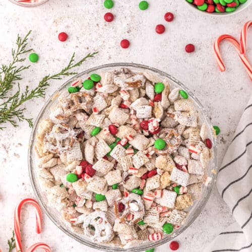 A bowl of reindeer chow with colorful M&Ms and peppermints on a white background surrounded by more Christmas candy