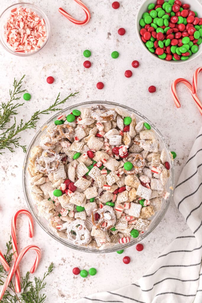 A bowl of reindeer chow with colorful M&Ms and peppermints on a white background surrounded by more Christmas candy