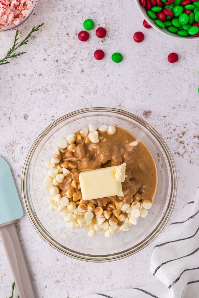 White chocolate chips, butter and peanut butter in a bowl next to a linen and a blue rubber spatula