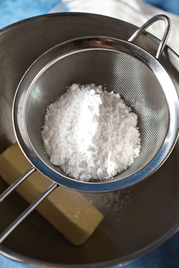 Powdered sugar in a sifter above a metal bowl with a stick of butter