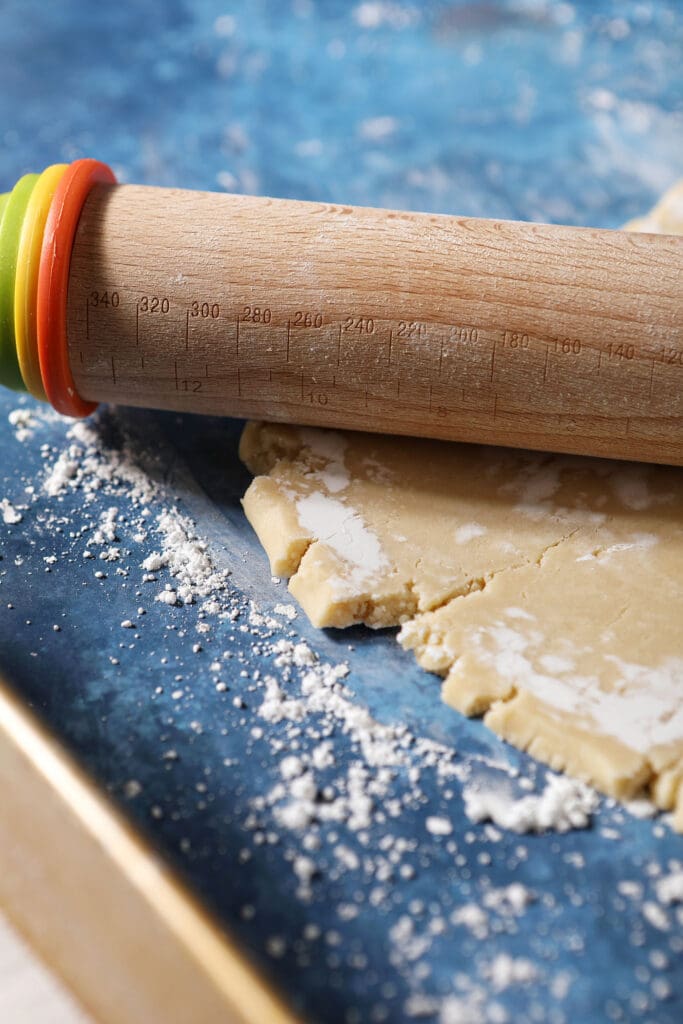 A wooden rolling pin rolls out cookie dough on a floured blue surface