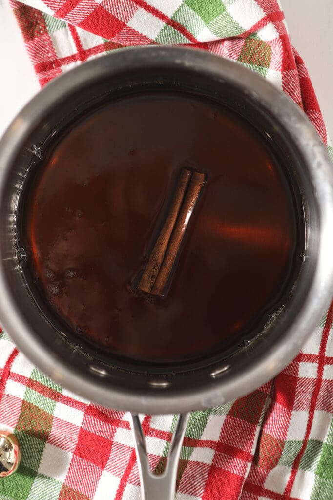 Maple syrup and a cinnamon stick together in a small saucepan over a red and green plaid towel