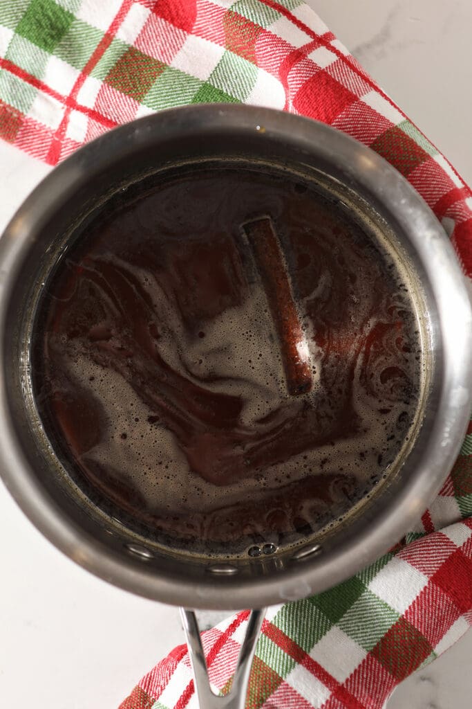 Hot maple syrup and a cinnamon stick together in a small saucepan over a red and green plaid towel