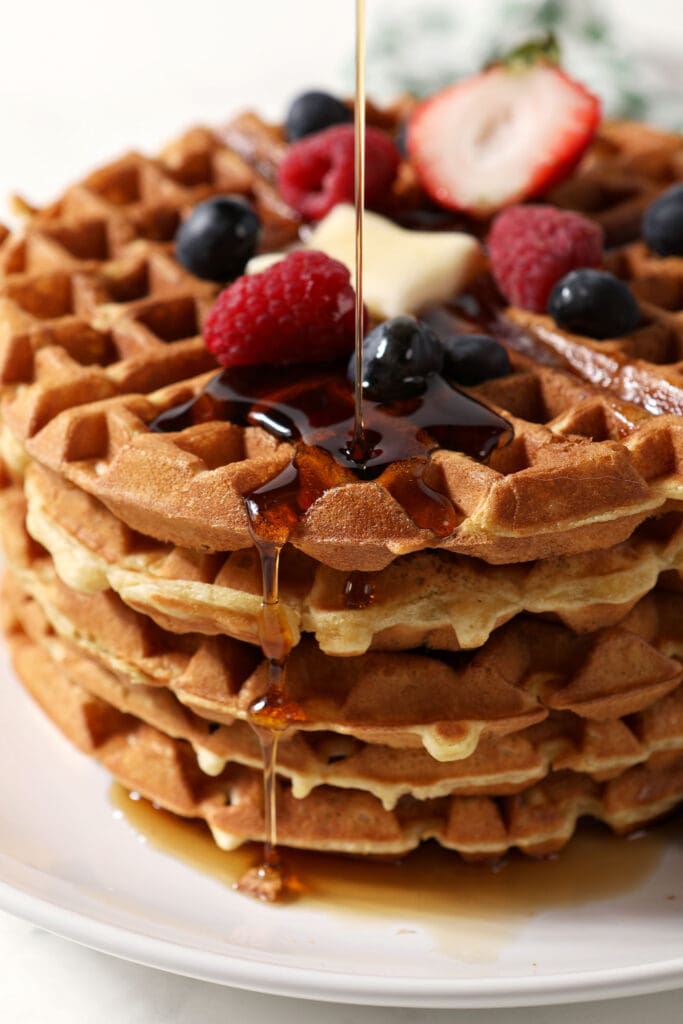 Syrup pours onto a stack of waffles on a white plate topped with fruit