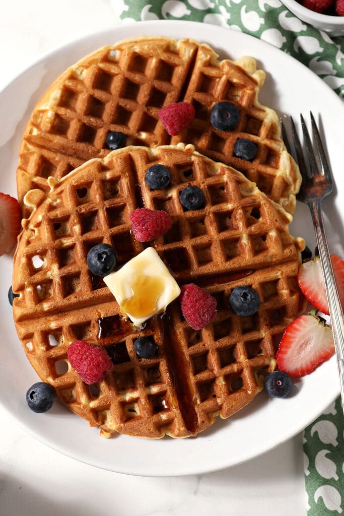 Two waffles on a white plate with a fork toped with berries, a pat of butter and syrup