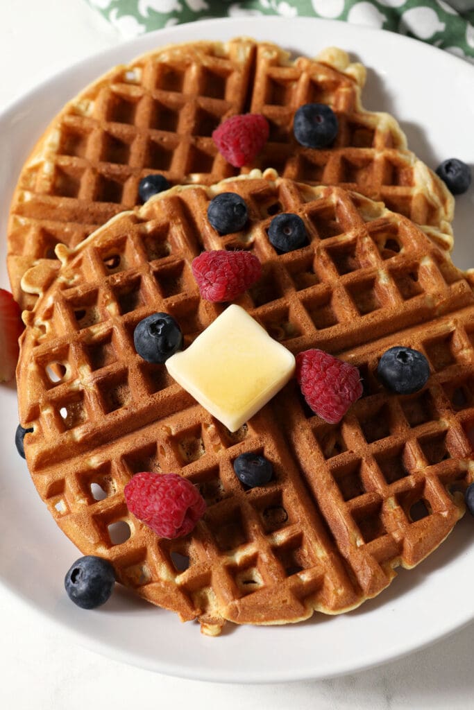 Close up of two round waffles on a white plate with a fork toped with berries and a pat of butter