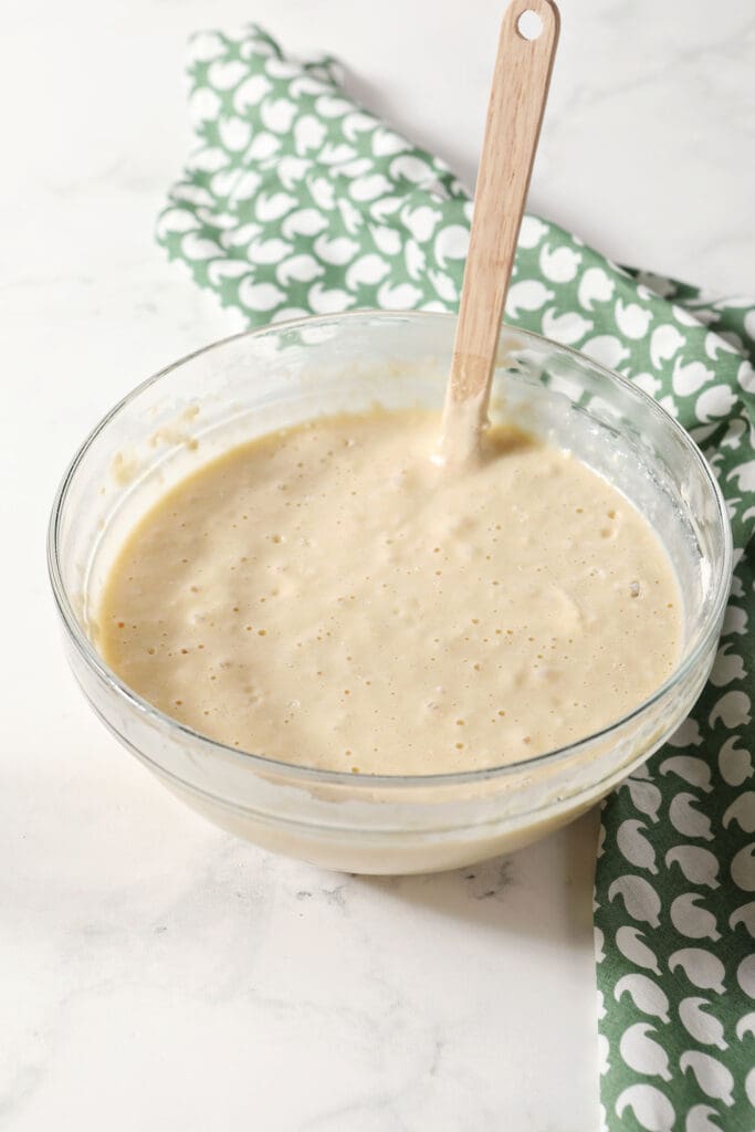 Waffle batter in a bowl with a rubber spatula on marble with a green patterned linen