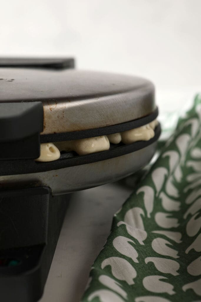 Close up of a waffle iron cooking waffles on marble next to a green patterned linen