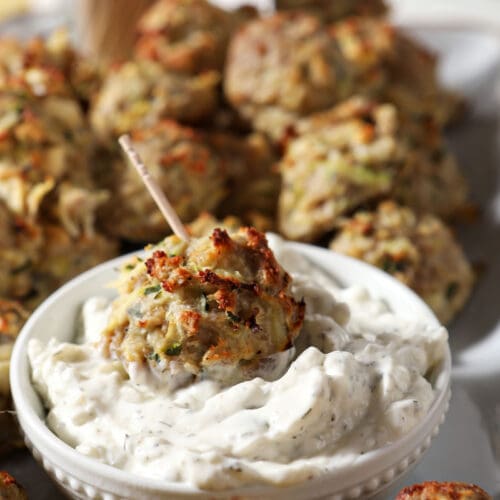 A Turkey Zucchini Meatball dipped in a bowl of tzatziki surrounded by more baked meatballs