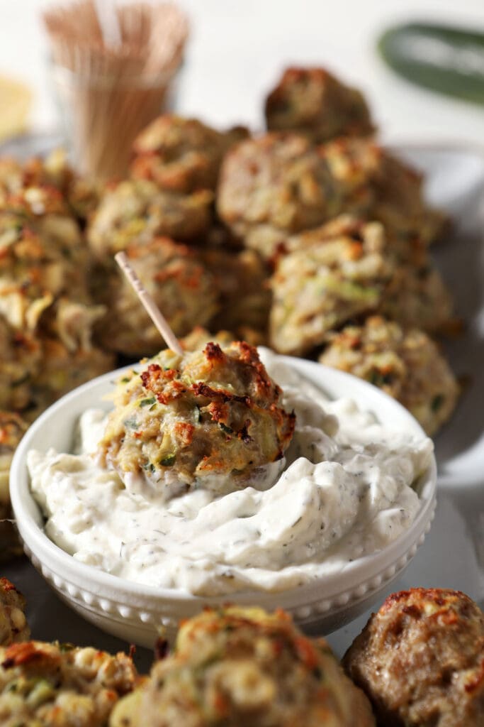 A Turkey Zucchini Meatball dipped in a bowl of tzatziki surrounded by more baked meatballs