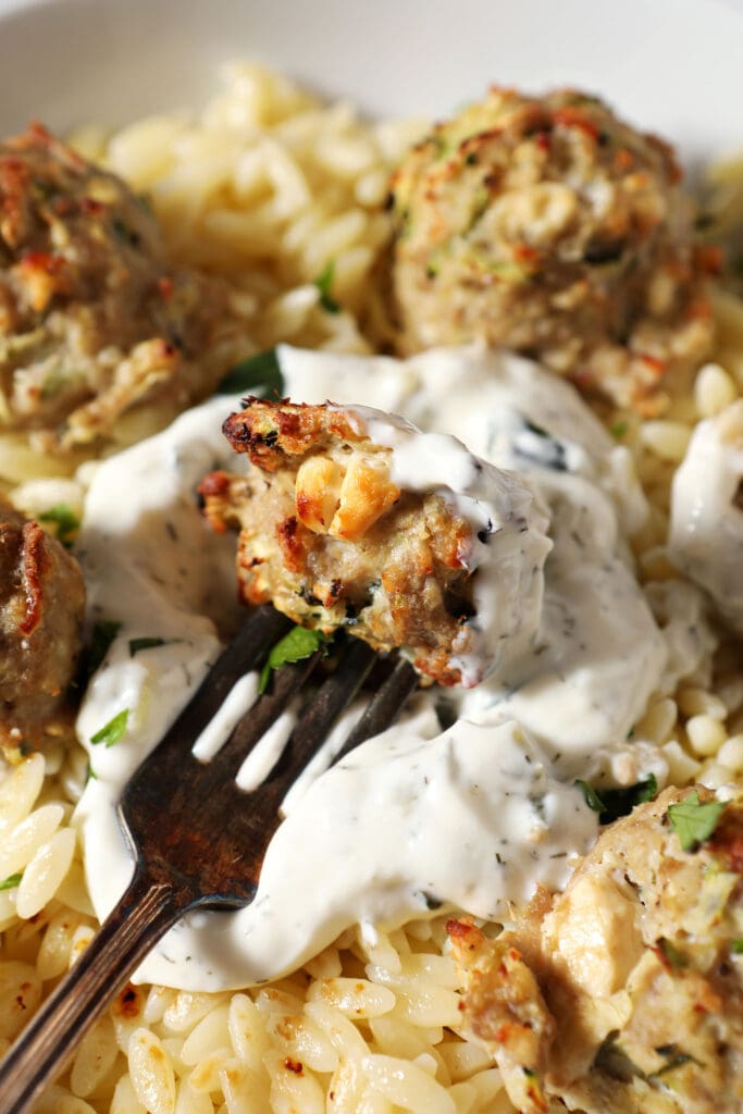 A zucchini meatball on a fork dips into a dollop of tzatziki in orzo pasta