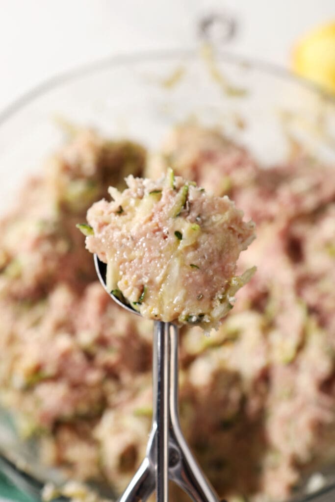 A cookie scoop holds a scoop of meatball mixture above a bowl