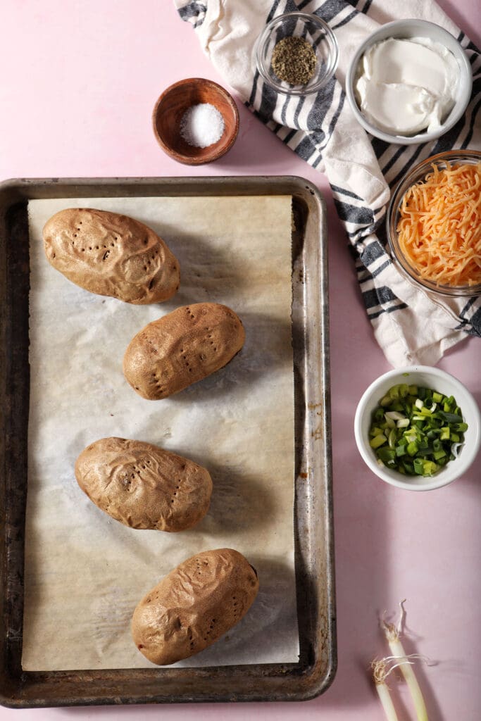 Baked potatoes on a sheet pan surrounded by twice baked potato ingredients