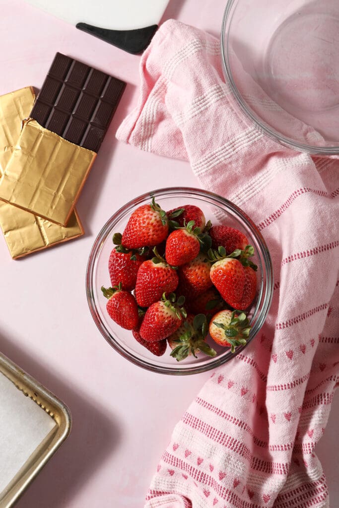 A bowl of strawberries next to bars of chocolate on a pink surface