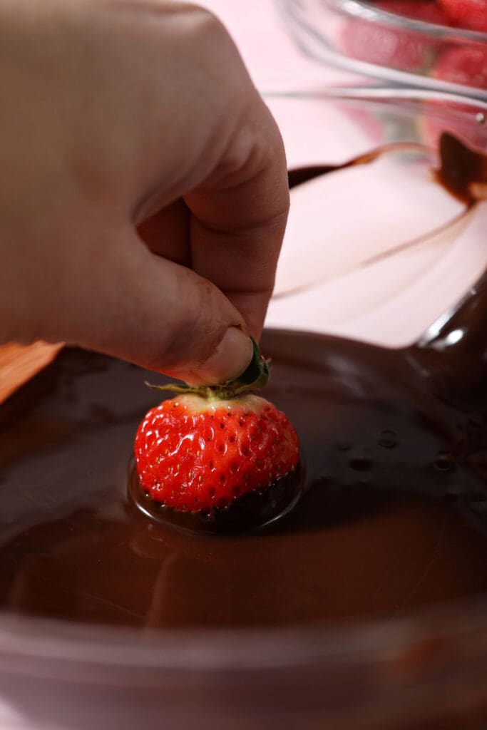 A hand dips a strawberry into melted chocolate