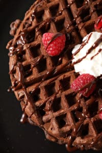 Overhead of a chocolate waffle drizzled with chocolate sauce, strawberries and whipped cream on a black plate